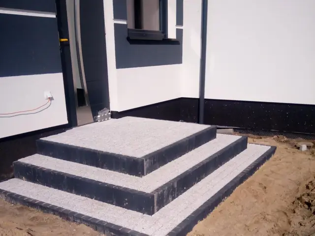 Trzy stopnie wejściowe z jasnej kostki z ciemnym obrzeżem przy drzwiach domu